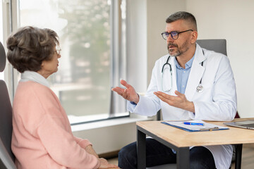 Middle aged professional male doctor talking while explaining medical treatment to senior female patient at consultation in clinic office