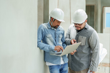 Two Asian male engineers with hard hats discussing project using laptop at construction site....