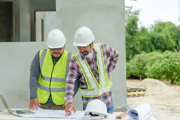 Two Asian male engineers with hard hats and safety vests reviewing blueprints at construction site....