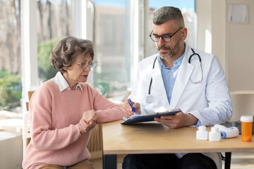 Man doctor in uniform with stethoscope holding documents, consulting older woman about checkup result, mature patient listening to physician explaining prescription