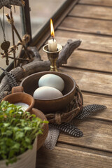 Atmospheric spring altar with burning candle, bowl with eggs, the first greens in the pot and birds feathers on wooden surface, vertical image, selected focus.