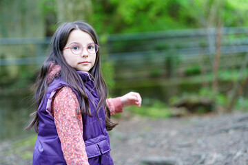 A little long-haired girl in glasses and a vest stands in the forest