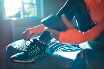 Active woman tying running shoes while holding smartphone in bedroom