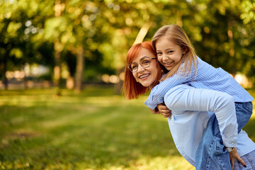 Fototapeta premium Lovely ginger-haired mother holding her young daughter on her back, smiling for the camera, enjoying nature together.