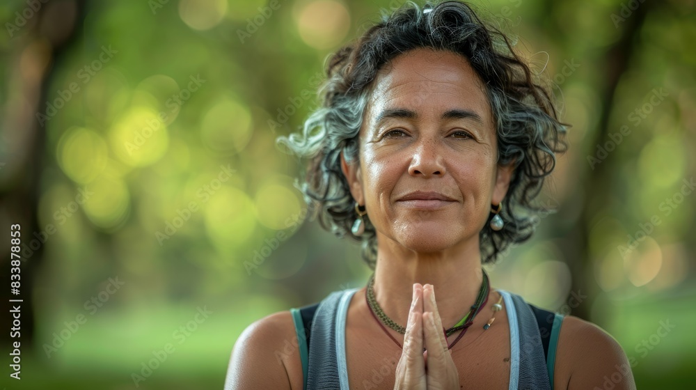 Wall mural A serene woman with curly hair wearing a necklace smiling gently with her hands clasped together in a prayer-like gesture standing amidst a lush blurred green forest.