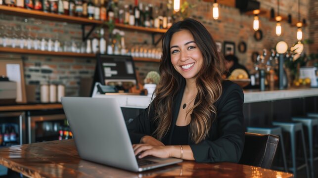 Happy young Latina businesswoman in suit working with laptop in a cafe or bar smiling confidently, for business owner, self employment, work from home, manager, small business, remote working concept.