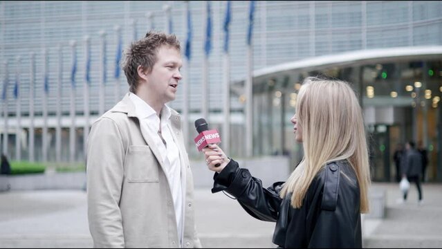A male interviewee and a female reporter engage in a discussion outside a modern office building. Professional setting with flags and glass entrance provide backdrop for their conversation