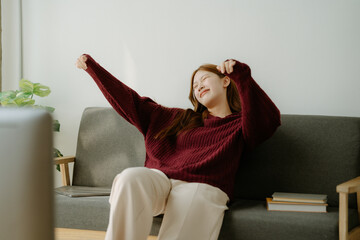 Young Asian woman feeling happy, smiling and watching TV while relaxing in the living room at home.