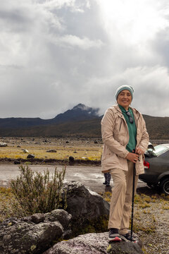 Full-body portrait of a Latina adult woman standing on a rock beside a dirt road with the sun behind her. She is wearing warm clothing on a cloudy and rainy day