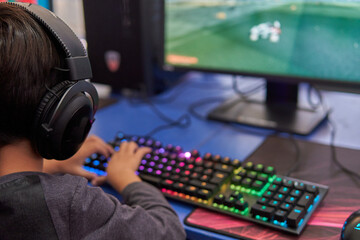 child with back headphones playing video games on a computer with a keyboard with lights.
