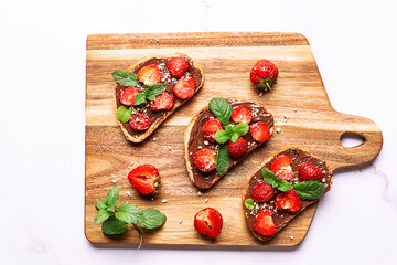 Sweet homemade toasts with chocolate paste, nuts, srtawberry and aromatic mint leaves on the serving plate on white stone background. Tasty summer dessert.