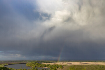 This image captures a dramatic sky over a tranquil river landscape, with a vibrant rainbow piercing through the storm clouds. The foreground features a meadow and farmland