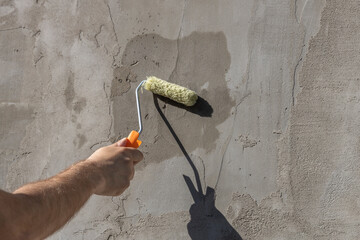 A worker applies the primer to the wall with a long-handled roller. A man's hand rolls a primer...