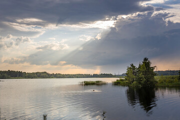 A person swims in a calm lake with a dramatic sky above. The sun shines through gaps in the clouds, casting a bright light on the water.