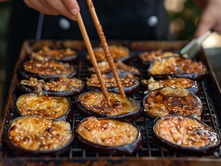 A person is using chopsticks to eat food that is on a grill. The food is made of eggplant and is covered in sesame seeds