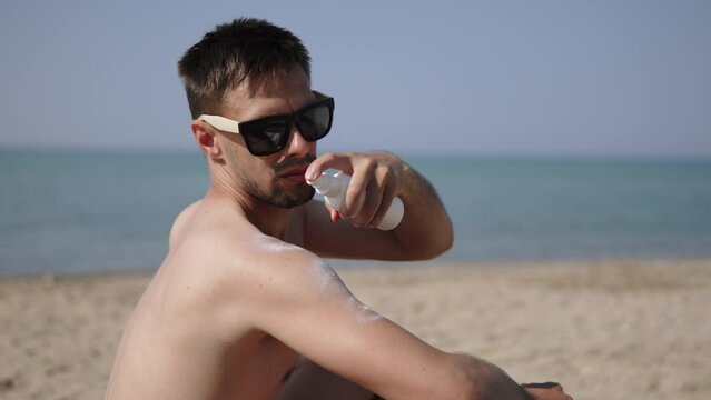 Young man applies sun protection lotion with spray for safe tan. Hispanic guy in sunglasses on sunny sandy beach of sea takes care of skin. Sunscreen protection, prevention of skin cancer.
