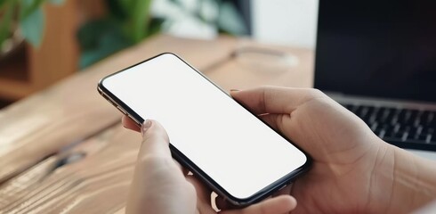 A closeup of hands holding an Phone with a blank screen, showing the phone's side view on a wooden desk.