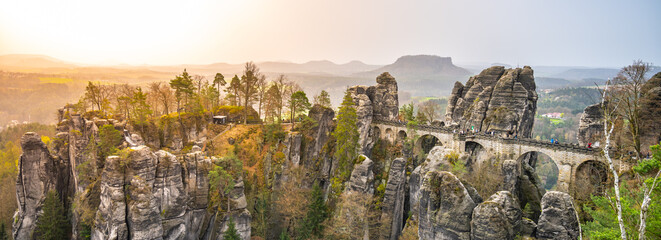 Golden sunlight bathes the Bastei Bridge and surrounding sandstone rock formations at dawn in Saxon...