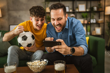 Two excited men friends brother use phone to watch football match home