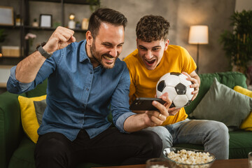 Two excited men friends brother use phone to watch football match home