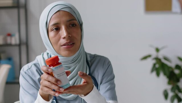 Medium close-up shot of smiling Muslim doctor in Islamic hijab giving jar of prescription medicine to anonymous dark-skinned male or female patient in GP clinic, warning about potential side effects