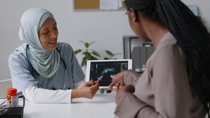 Medium shot of smiling female gynaecologist in Islamic hijab sitting in clinic, holding digital tablet, demonstrating video of foetus ultrasound to pregnant black woman, pointing, answering questions