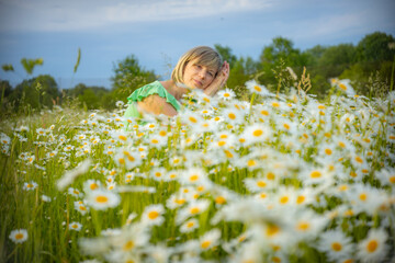 Fototapeta premium A woman in a green shirt rests her head on her hands in a field of blooming white daisies with yellow centers. The background features lush greenery and a clear sky, creating a serene and peaceful sce