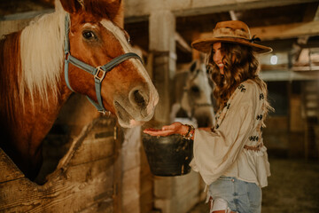 Smiling cowgirl feeding a horse in rural stable at her ranch.