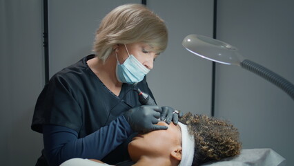 Cosmetician tattooing permanent eyebrows in beauty studio. Woman holding machine