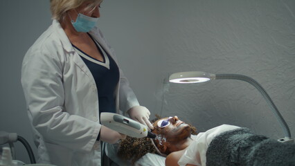 Dermatologist doing facial peeling with carbon mask. Woman lying with glasses