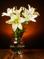 A vase of white lily flowers sits on a wooden table. The flowers are in full bloom. The vase is...