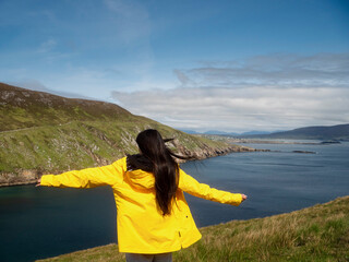 A teenager girl in a yellow jacket is looking out over a green hillside with a beach and ocean in...