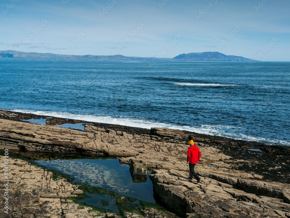 Wall mural a man in a red jacket walks on a rocky beach looking out at the ocean. the sky is clear and the wate