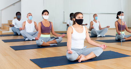 Fototapeta na wymiar Multiethnic group of young adults in face masks for viral protection sitting in lotus position practicing meditation at yoga class