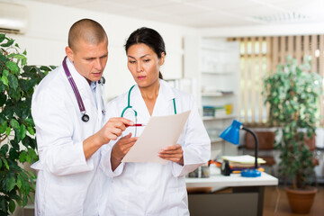 Two professional doctors standing in office, reading medical card of patient and discussing diagnosis..