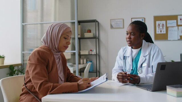 Medium shot of young Middle Eastern Muslim patient in hijab sitting in GP clinic during appointment with physician, filling out insurance paperwork, and black female doctor explaining what to write