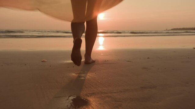 Woman in dress fluttering in wind walks barefoot on sandy beach of ocean towards sun at sunset.
