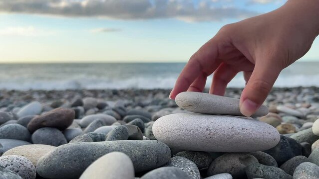 4K. Close up female hand builds pyramid of gray stones on pebble beach with sea waves. White woman experiences zen on ocean shore. Spa, calmness, yoga, self-improvement, construction, games, travel
