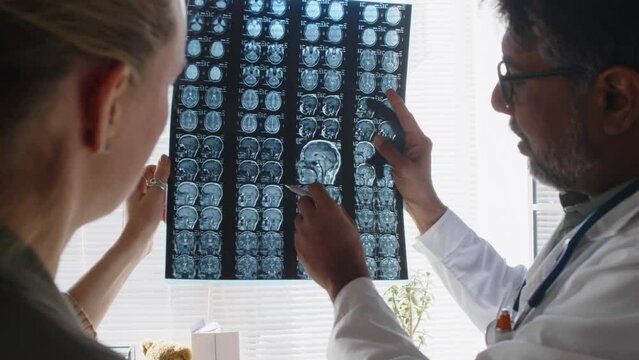 Medium rear shot of mature Indian male neurologist demonstrating Caucasian female patient MRI scan and pointing abnormalities after brain injury, while consulting in medical clinic