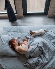 Top view of a red-haired Caucasian woman lying in bed with her baby son and Jack Russell terrier...