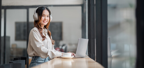 A woman wearing headphones is sitting at a table with a laptop and a cup of coffee. She is focused...