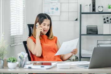 A woman in an orange shirt is talking on her cell phone while reading a piece of paper. The scene is set in a home office with a desk, a chair, a laptop, and a potted plant