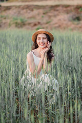 A woman is sitting in a field of tall grass, wearing a straw hat and a white dress. She is smiling and she is enjoying the outdoors