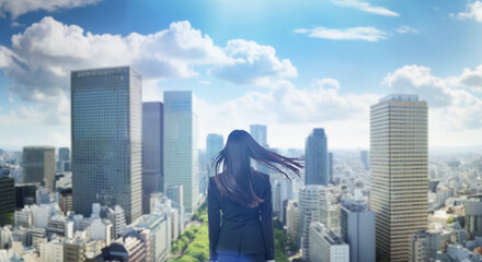 日本の街・都市・朝日を受けたオフィスビル群を前に佇み風を受けて髪をなびかせる会社員の女性の出勤　就職・ビジネス・はじまり・決意・目標・挑戦のイメージ
