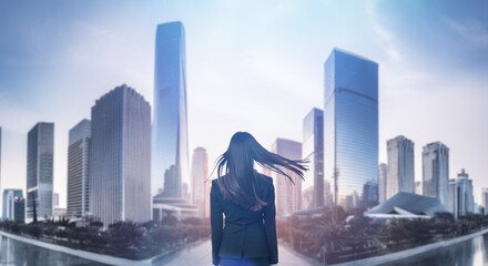 日本の街・都市・朝日を受けたオフィスビル群を前に佇み風を受けて髪をなびかせる会社員の女性の出勤　就職・ビジネス・はじまり・決意・目標・挑戦のイメージ