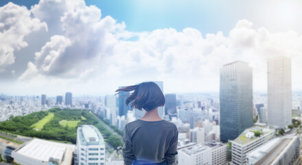 日本の街・都市・朝日を受けたオフィスビル群を前に佇み風を受けて髪をなびかせる会社員の女性の出勤　就職・ビジネス・はじまり・決意・目標・挑戦のイメージ