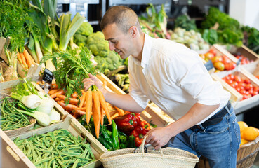 Attentive young man purchaser holding organic carrots at the counter in large supermarket