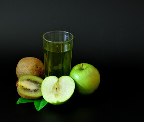 A tall glass of freshly squeezed fruit juice on a black background, next to a kiwi fruit and a ripe green apple.