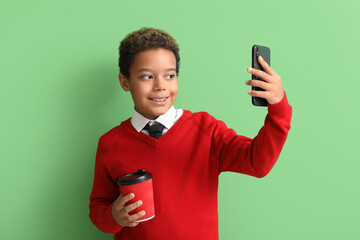 Little African-American boy with cup of coffee taking selfie on green background