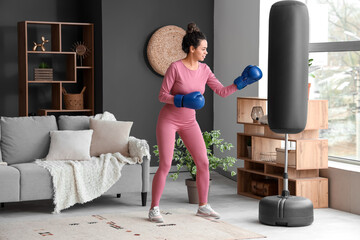 Sporty young African-American woman with boxing gloves punching bag in living room